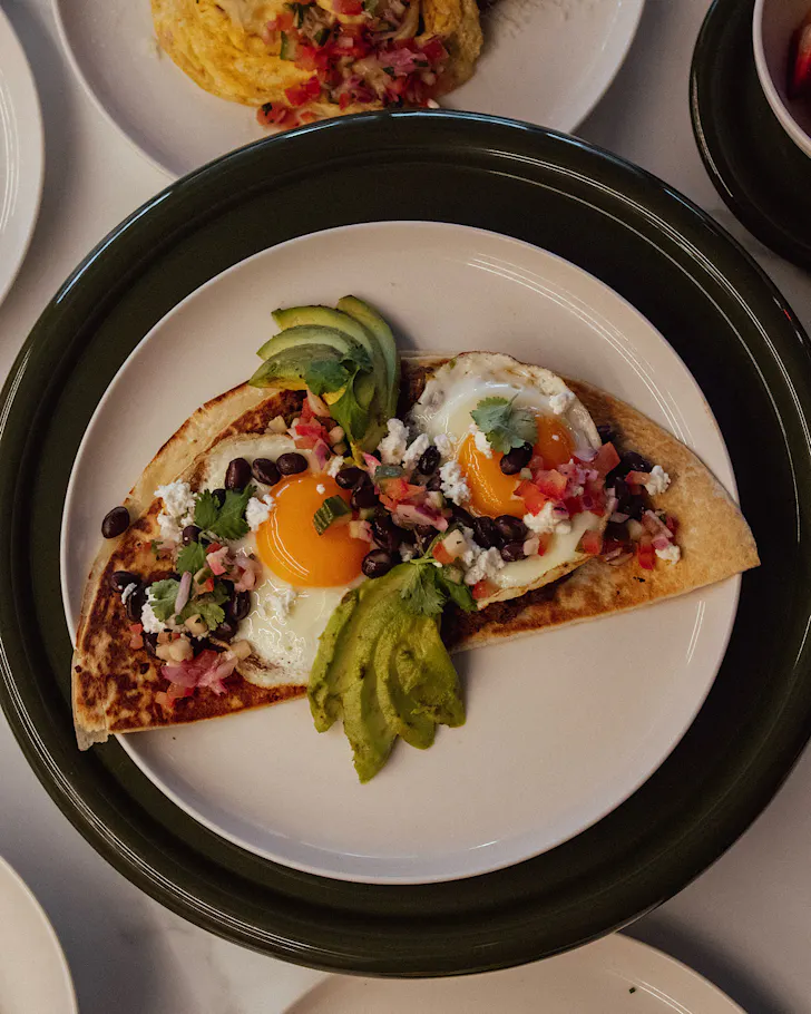 A breakfast quesadilla topped with two fried eggs, beans and coriander, and avo on a white plate.