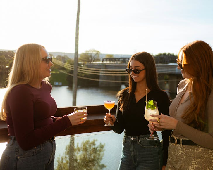 Three women chat and smile on the balcony of Sinclair's while holding drinks in hand. The sun is setting and the Nepean River is in view behind them.