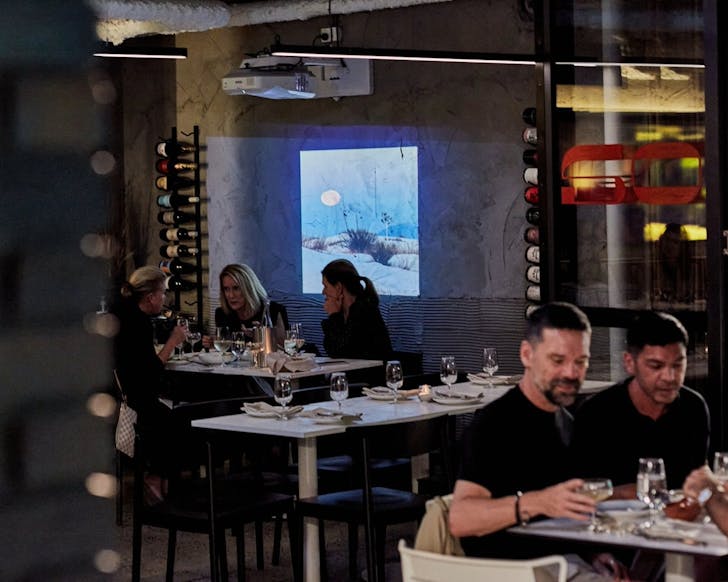 Indoor seating inside Sonora. Groups of people sit at different tables inside a modern, sleek and low-lit restaurant. A projection of artistic visuals is played on one of the dark walls.