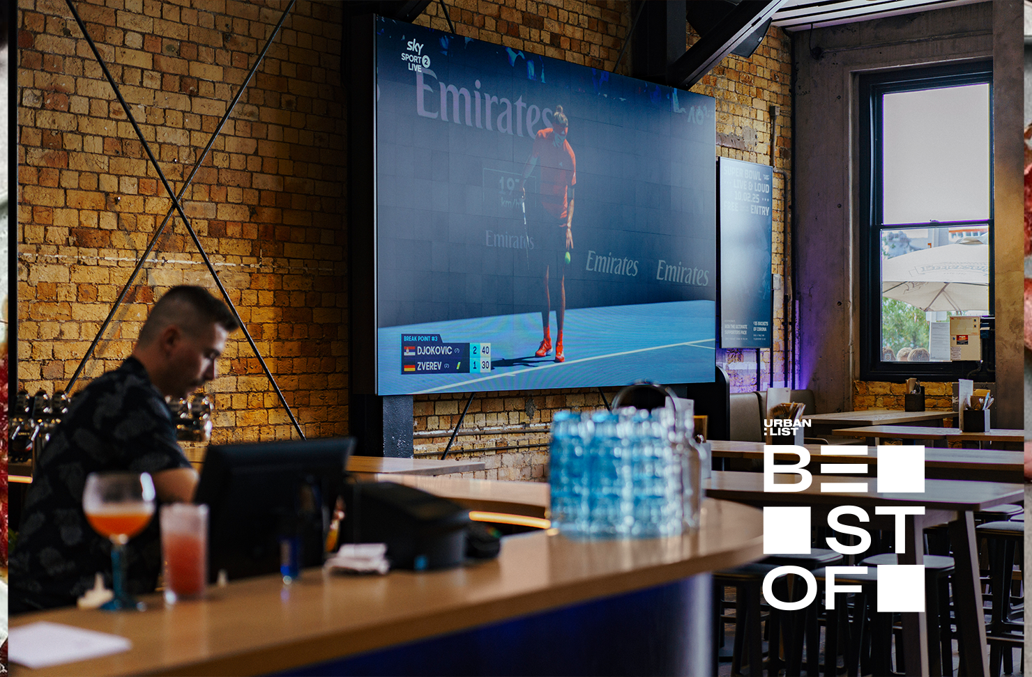 A bartender is busy behind the bar while a sports game plays on a massive screen at Tyler St Sport. 
