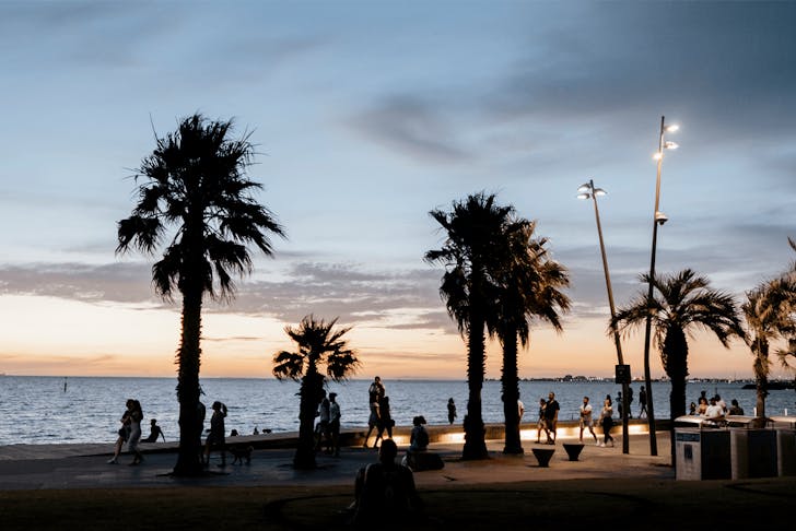 A sunset looking over the beach with palm trees and people walking along the foreshore. 