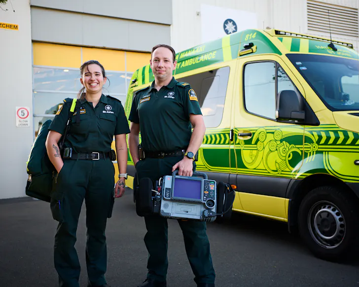 Two St John staff stand in front of a yellow and green ambulance. 
