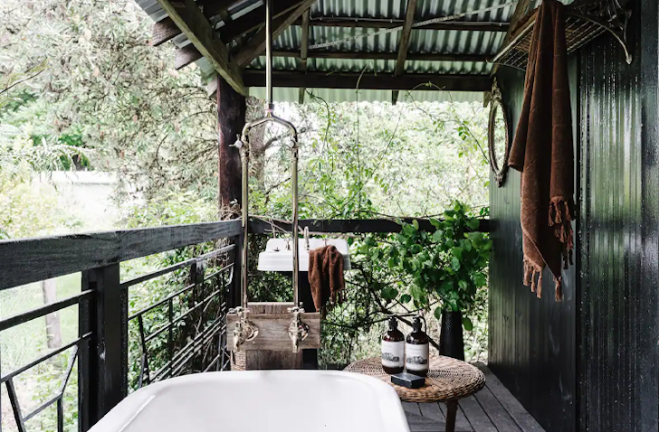 A white free-standing bathtub sits on an undercover balcony next to a table with bath soap.