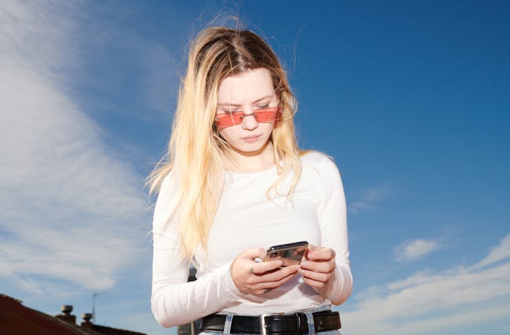 A blonde woman wearing a pink top and glasses glances down at her phone. 