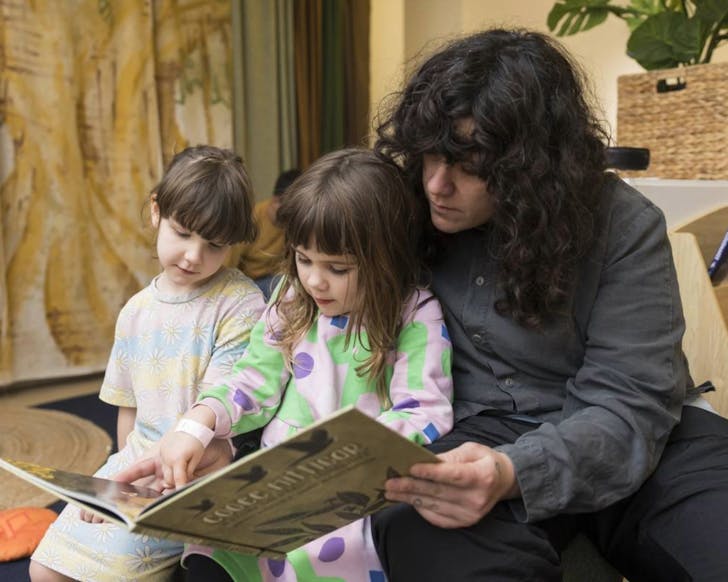 A parent sits with two children, while all three of them look down at a story book that is open in front of them. They are reading peacefully together. 