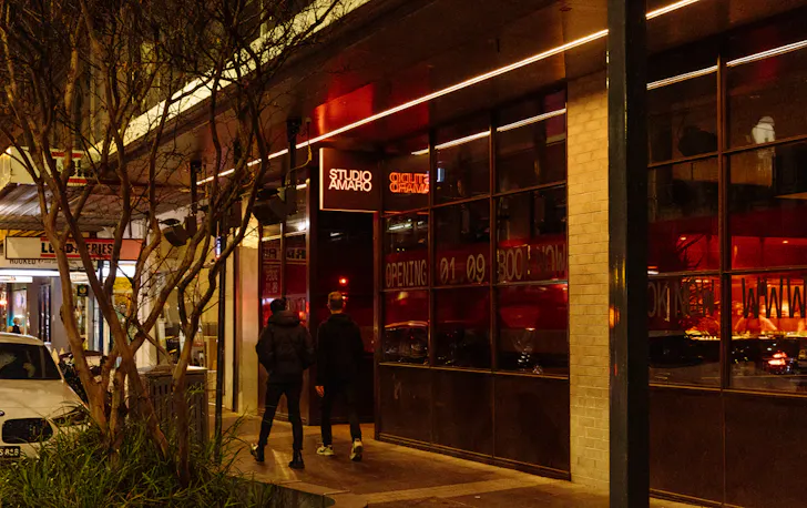Two people walking past a lit up restaurant. 