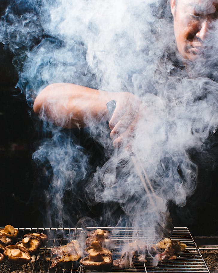 A chef is almost entirely obscured by smoke from chargrilling mushrooms. 