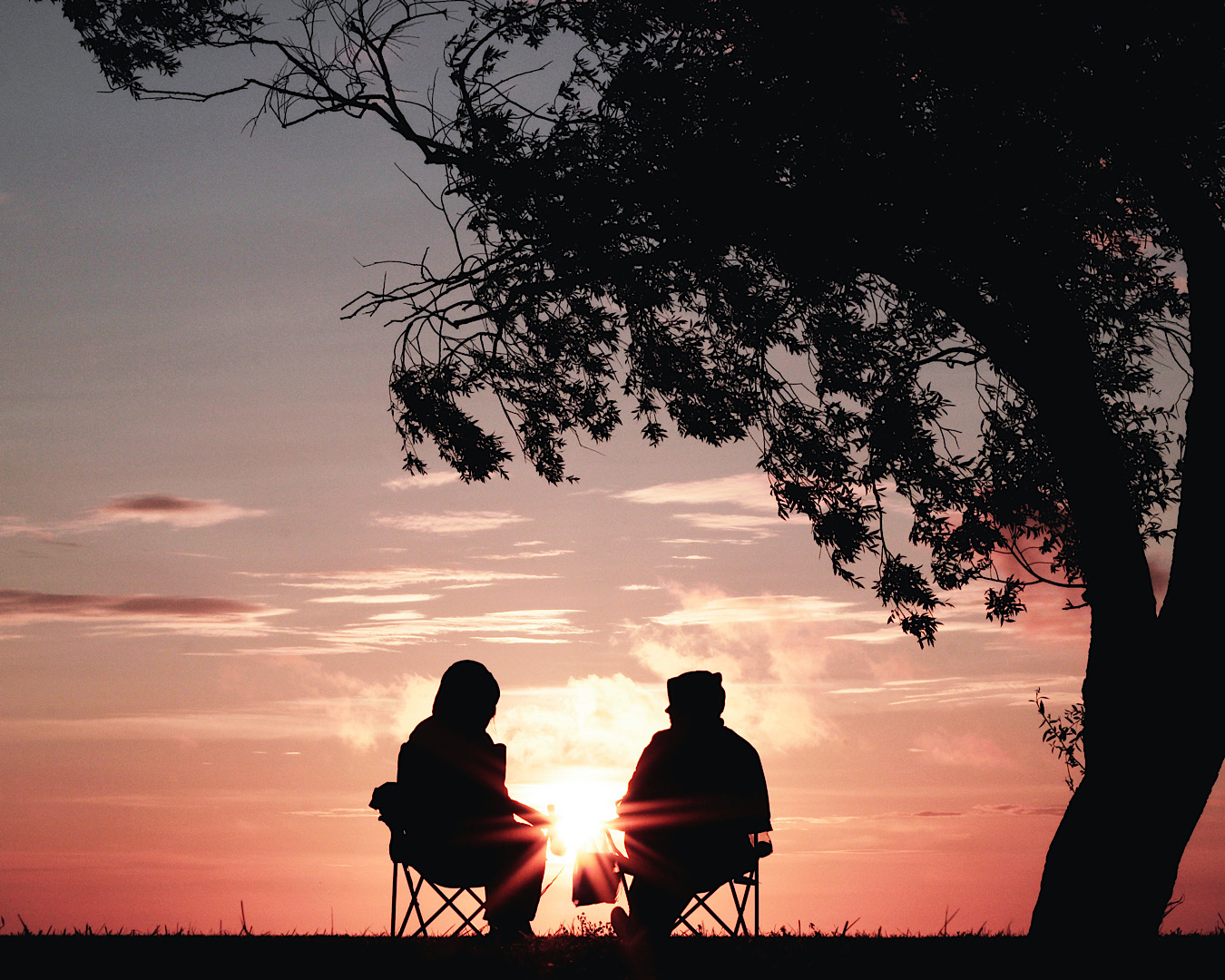 Two friends sit on fold up chairs on the top of a hill watching the sunset. The light has a silhouette effect. 