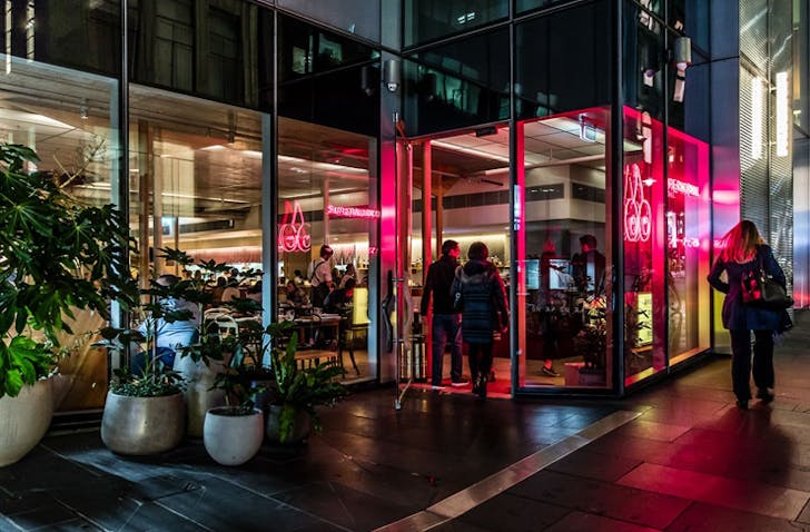 The outside of a busy restaurant with people dining and neon lights.