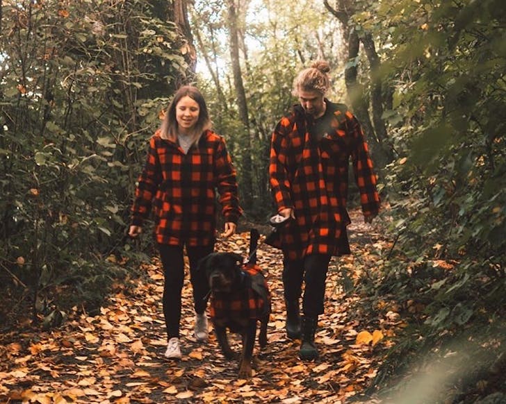 Two people walk a dog through the woods all wearing matching red and black Swanndri hoodies. 