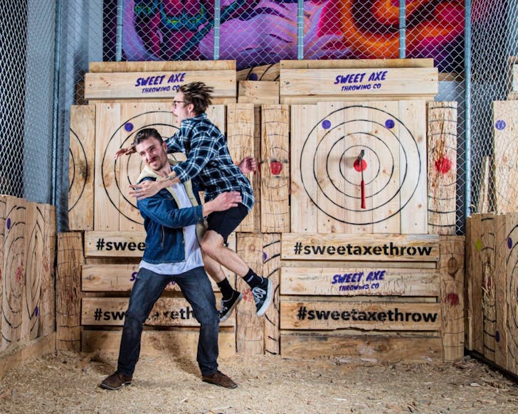 Two men have just thrown axes. One has scored a bullseye and is jumping in the air in celebration. 