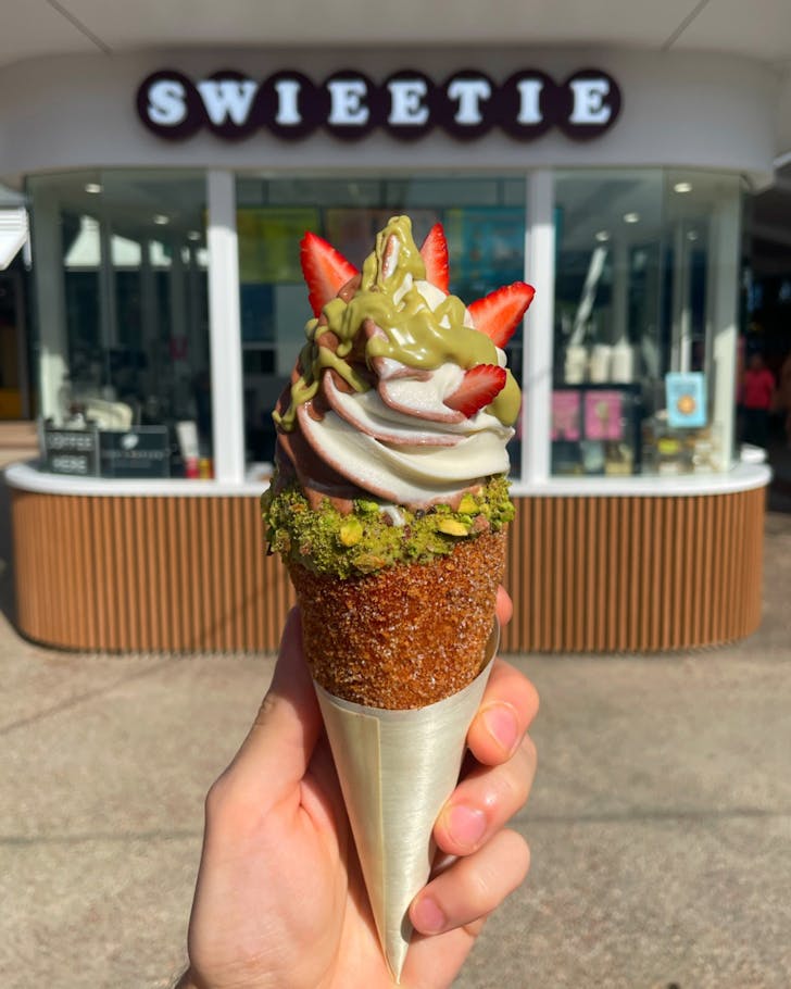 Chimney cake pastry filled with vanilla and chocolate soft serve, topped with pistachio pieces and strawberries, being held up in front of Swieetie shop