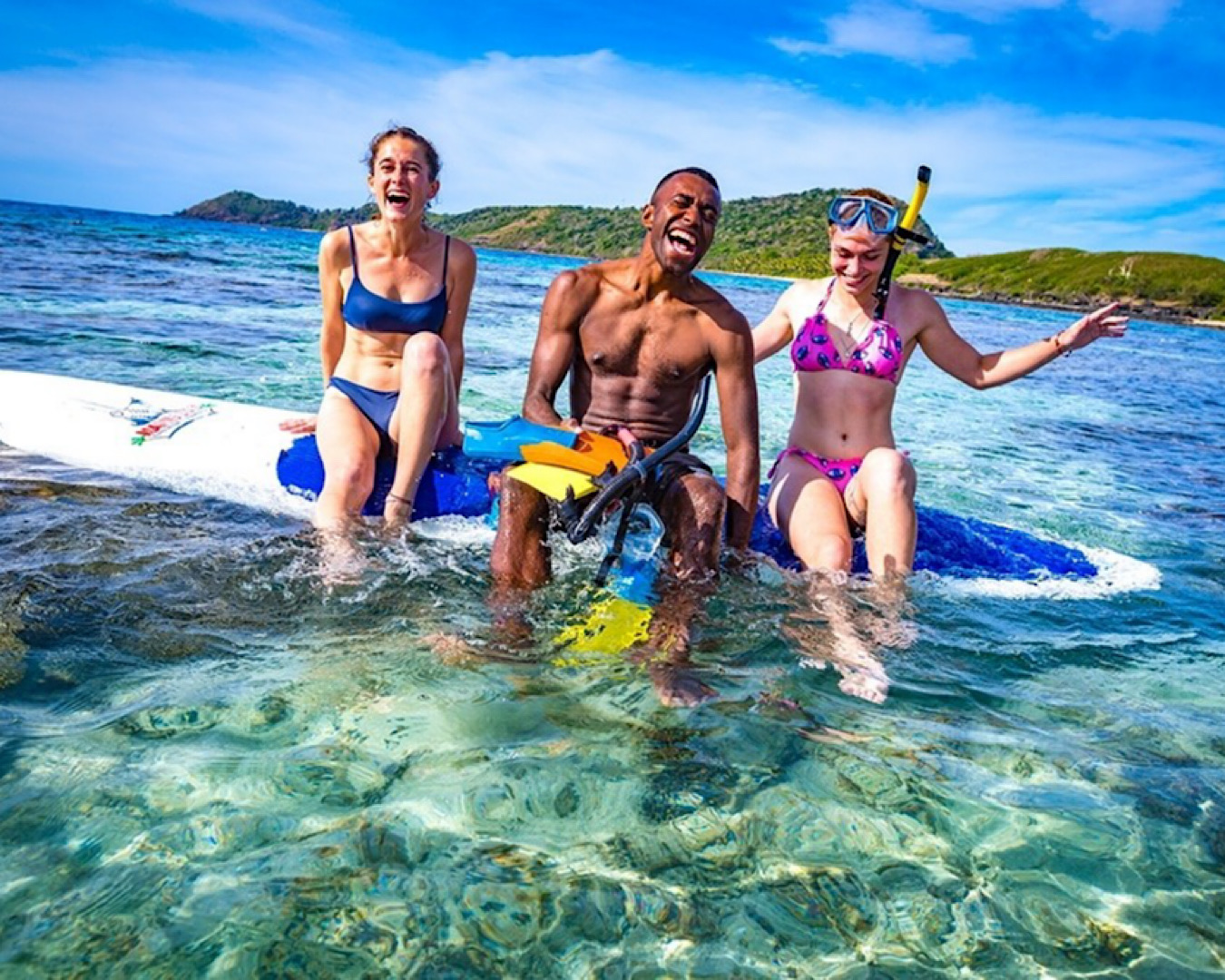 Two women wearing bikinis and one man holding snorkelling gear float on a paddle board at the Yasawa Islands. 