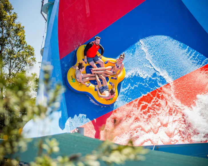 A wide shot of four people on a floating yellow water seat as they go sideways on a water ride. They have smiles on their faces and in the foreground there is some greenery, with the bright sky peaking through behind the water ride.