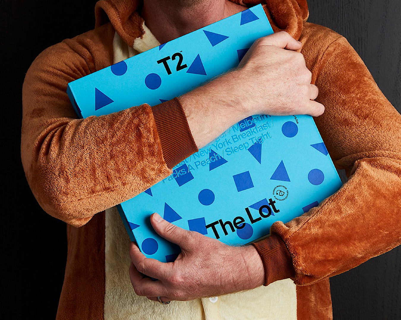 A dad in a comfy-looking brown jumper hugs a bright blue box of T2 The Lot with glee after being gifted it on Father’s Day. 