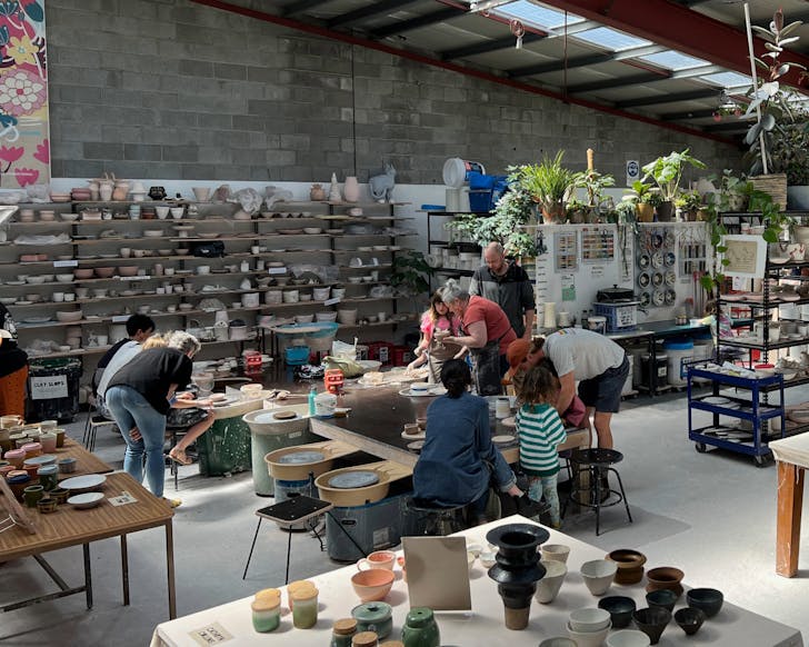 A wide shot of inside a large ceramic studio where there are tables and shelves stacked with ceramic decor. In the centre of the room a group of several people of all ages are bent around a table of ceramics and working with some clay.