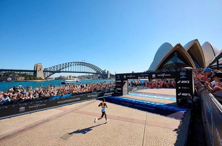Wide view image of the finishing spot of the TCS Sydney Marathon. It is set up at the Sydney Opera House Forecourt, and there is the view of spectators waiting on the sidelines and the Sydney harbour and Harbour Bridge in the background. 