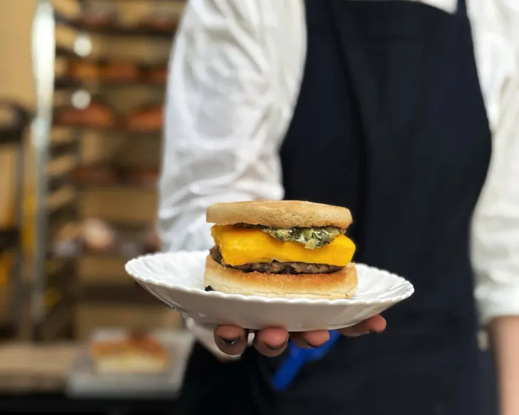 Chef hand holding out plate of egg, bacon and cheese sandwich