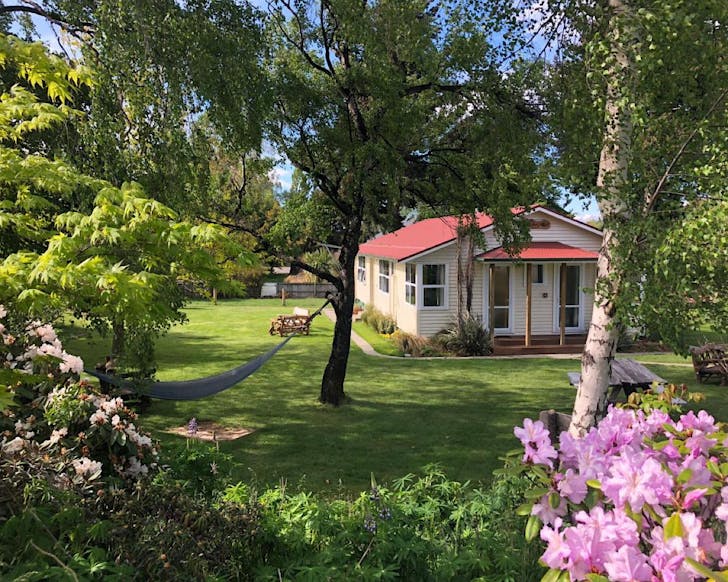 A cute cottage sits on a joyful lawn peppered with trees. A hammock hangs from two of the trees. 