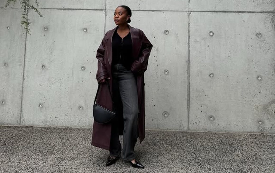 Woman dressed in black with maroon trench coat posing in front of concrete wall