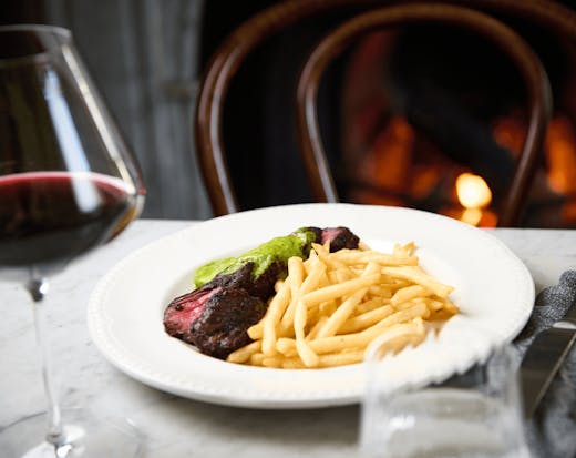 A plate of steak frittes with a glass of wine and a fireplace. 