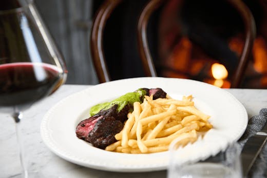 A plate of steak frittes with a glass of wine and a fireplace.