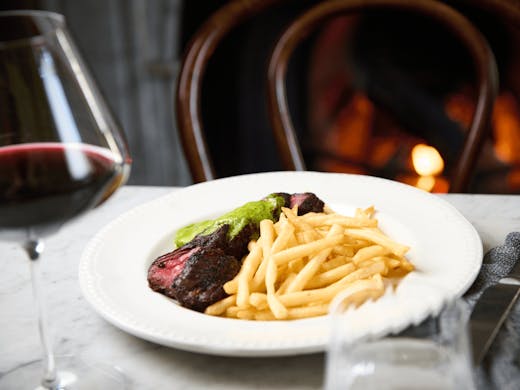 A plate of steak frittes with a glass of wine and a fireplace. 