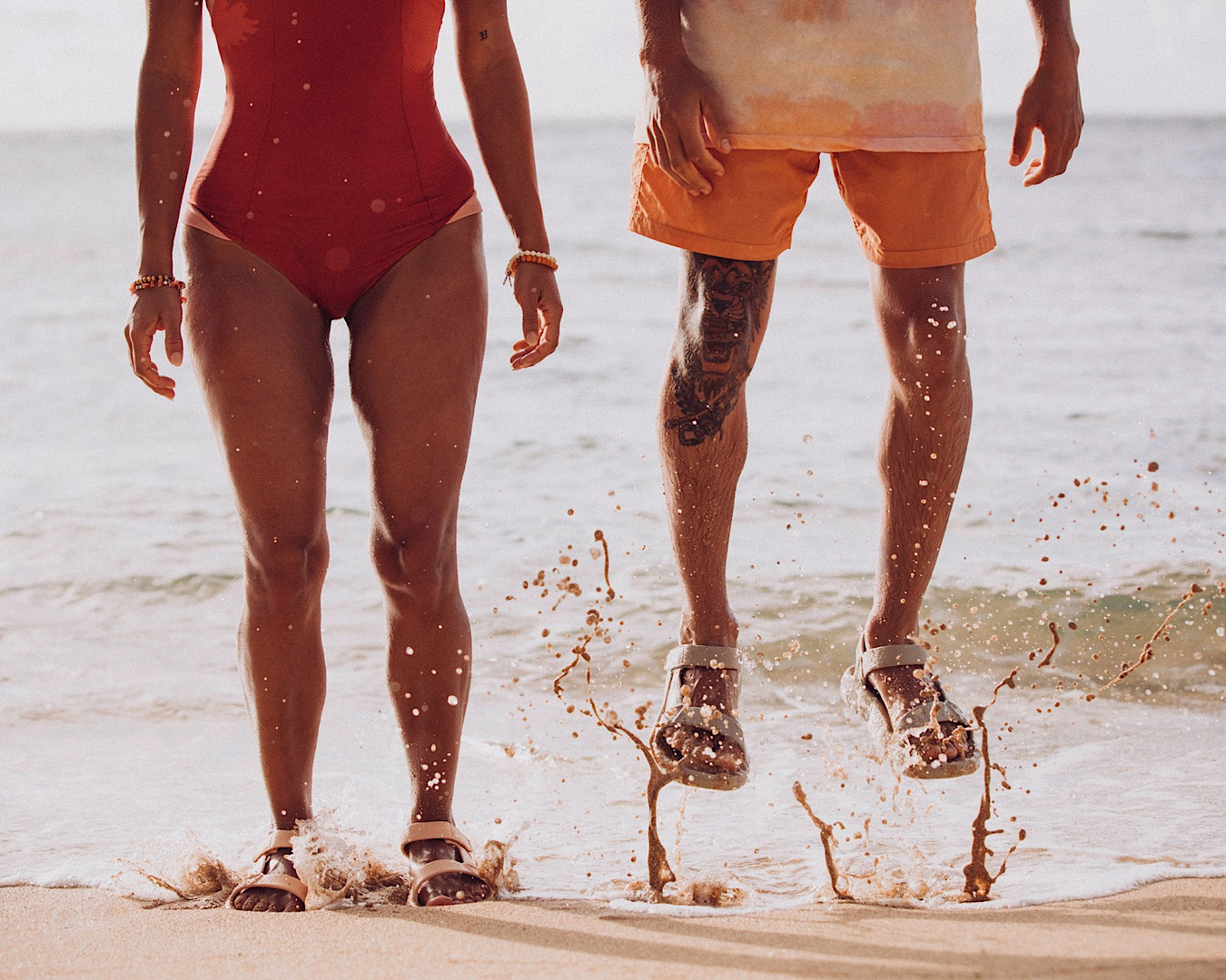 Two people jump in the shallow waves on a beach, wearing Tevas. 