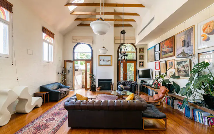 A chesterfield leather couch sits in the centre of a Airbnb Melbourne living room. Above it, exposed timbers line the ceiling, and next to it, a book shelf packed with coloured books and indoor plants.