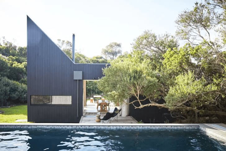 An infinity pool looking out onto an angular black house with trees covering part of the roof. 