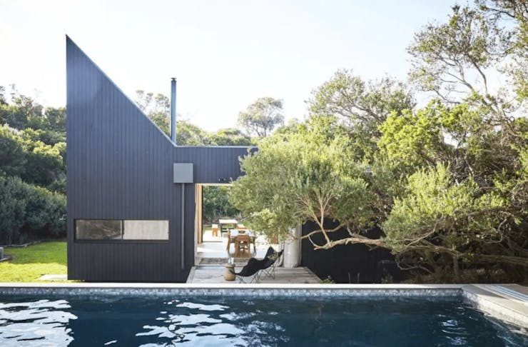 An infinity pool looking out onto an angular black house with trees covering part of the roof. 