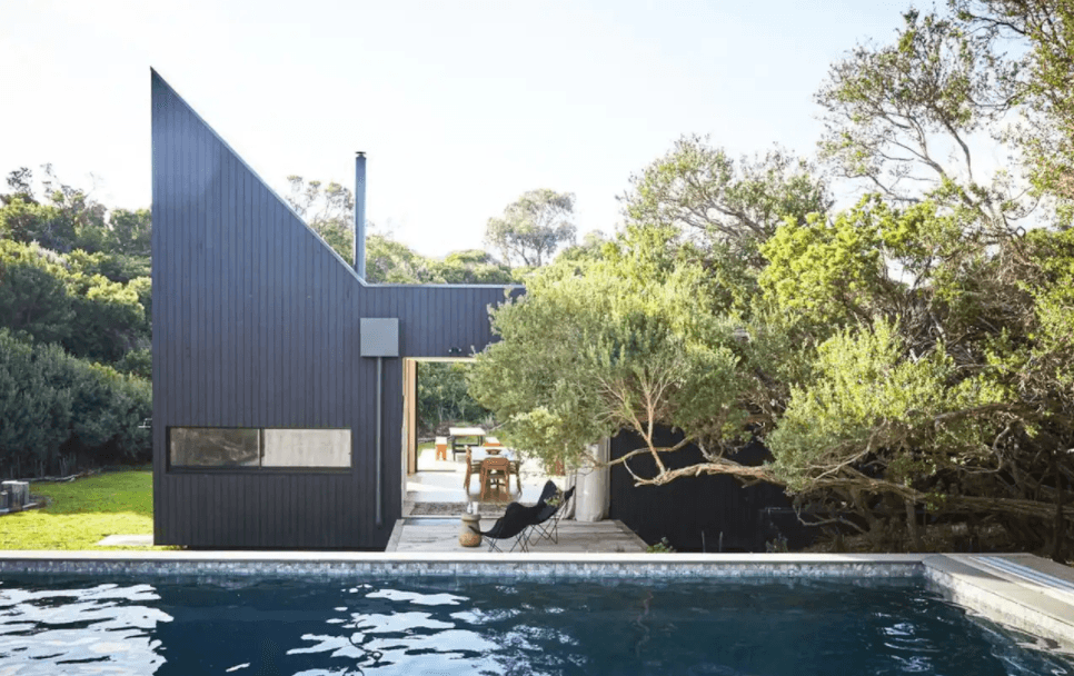An infinity pool looking out onto an angular black house with trees covering part of the roof. 