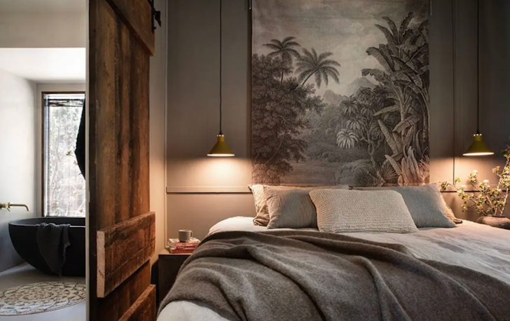 A queen-sized bed with two lamps on either side and a bath can be seen on the other side of the partial wall.