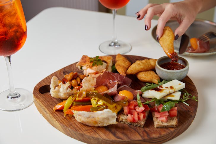 A hand reaches for food from a platter of charcuterie, next to two Aperol Spritz. 