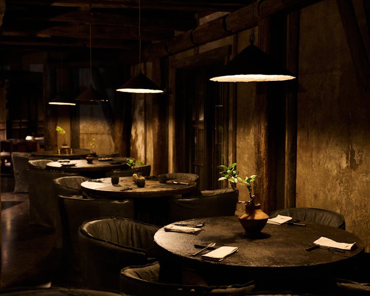 A dimly lit room with dark interiors and low-hanging light shades over dark, round tables set with cutlery and clay crockery.