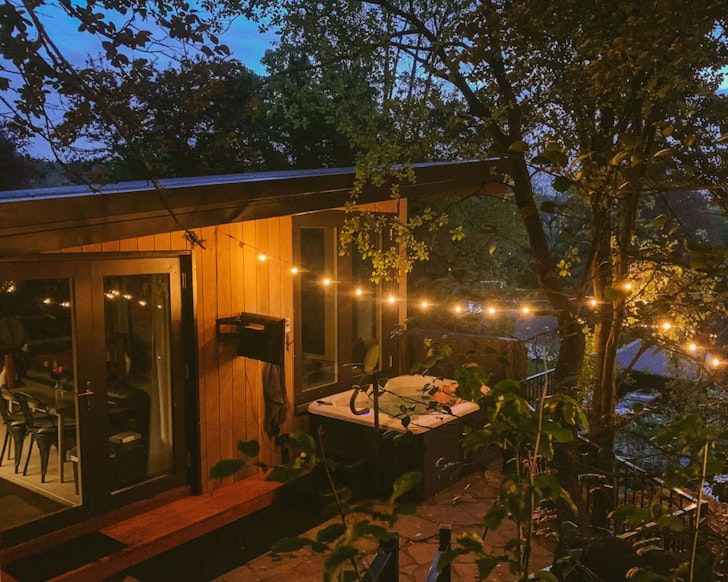 An exterior view of The Bird's Nest romantic stay in New Zealand with hot tub and festoon lights outside