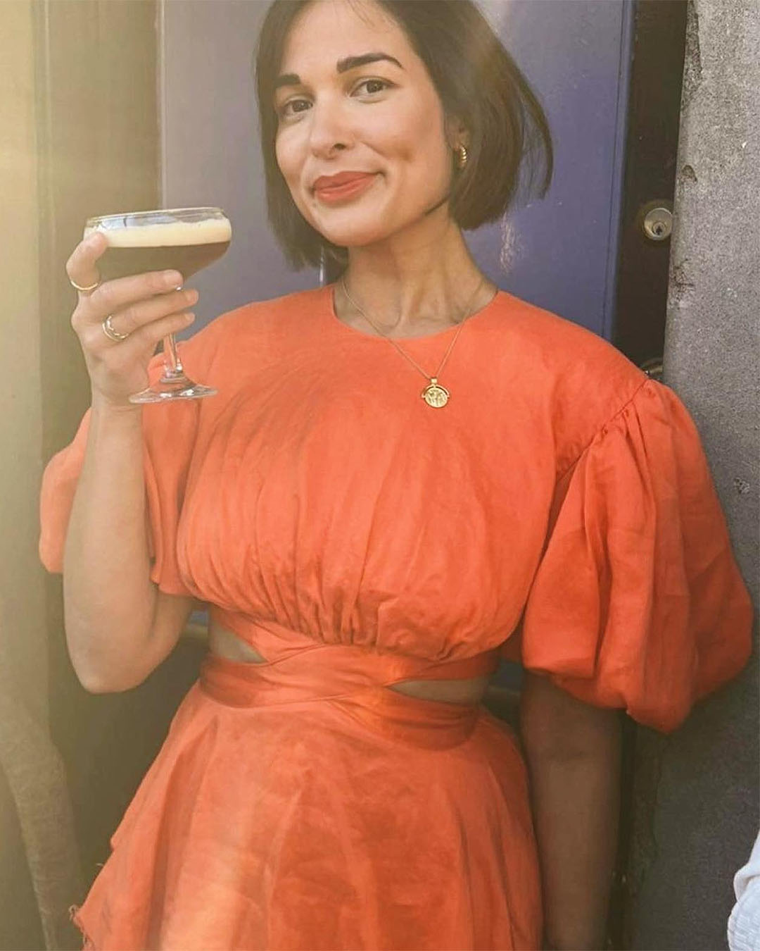 A woman in a beautiful orange dress holds an espresso martini while looking into the camera.  She is wearing a dress available to rent from HireSociety, one of New Zealand's best online dress hire shops.