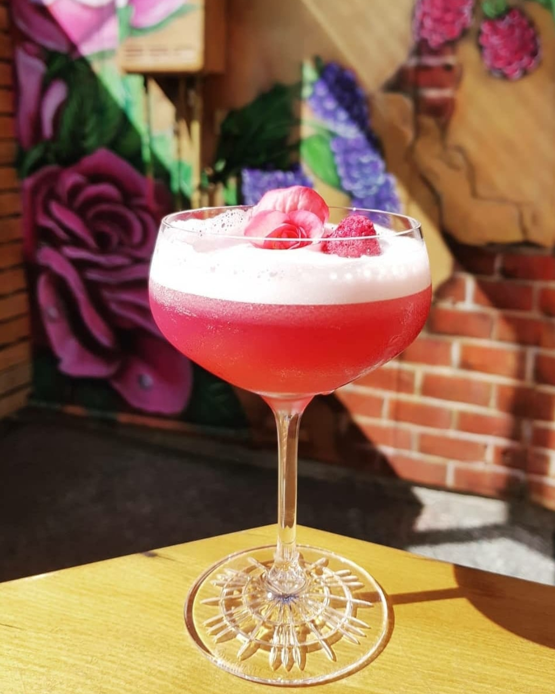 A delicious-looking, vibrant pink cocktail garnished with petals and berries sits on a table, ready to be sipped. 