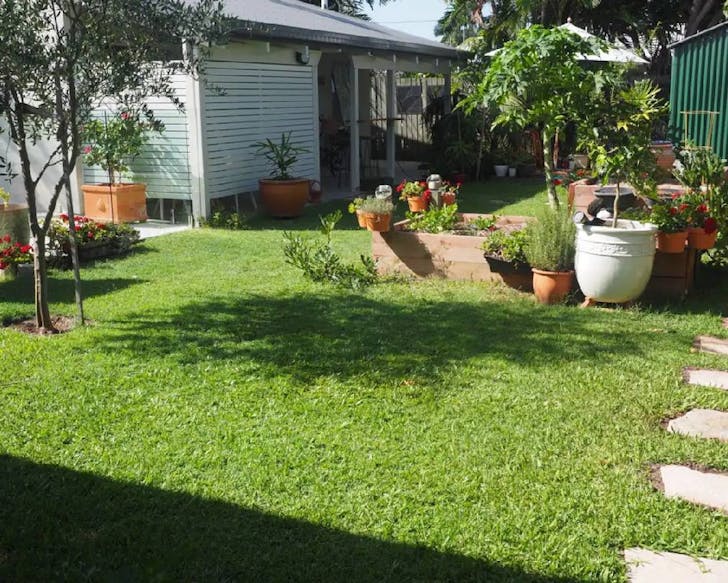 Outdoor garden area on a bright sunny day. Well mown bright green grassy area with potted pink/red flowers and leafy green plants. Cottage with a white privacy screen can be seen in the left side of the image. There is a 'stepping stone' path to the right of the garden and a few sparsely spaced, small trees. The Cottage is a perfect play to stay next time you're in Maroochydore. 