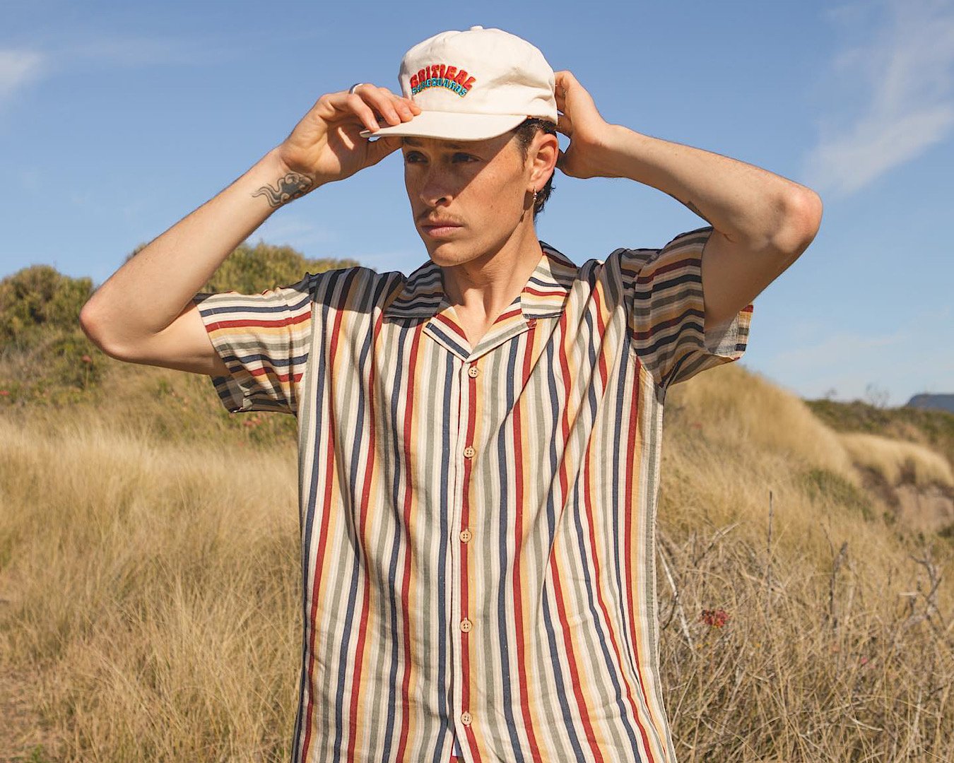 A young man standing outside wears a stripy shirt and a white snapback cap with the words ‘Critical Surfboards’ embroidered on it in orange and light blue. 