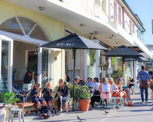 Outdoor view of The Depot. Many people are sitting in outdoor seating around the cafe, the weather is bright and nice and the outside of the cafe building is painted in colourful pastels of yellow, pink and blue. 