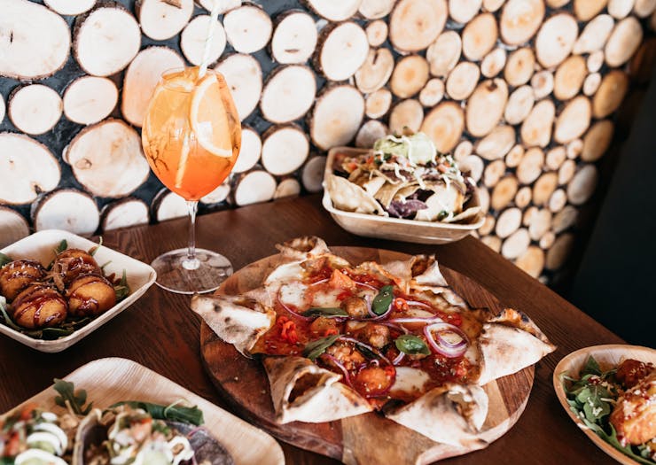 A table filled with cocktails, pizza, tacos and arancini balls behind a backdrop of a wooden wall.