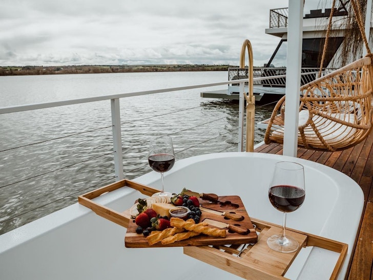 An outdoor bath, set with a cheesboard and glasses on red wine, sit on the deck of a floating home.