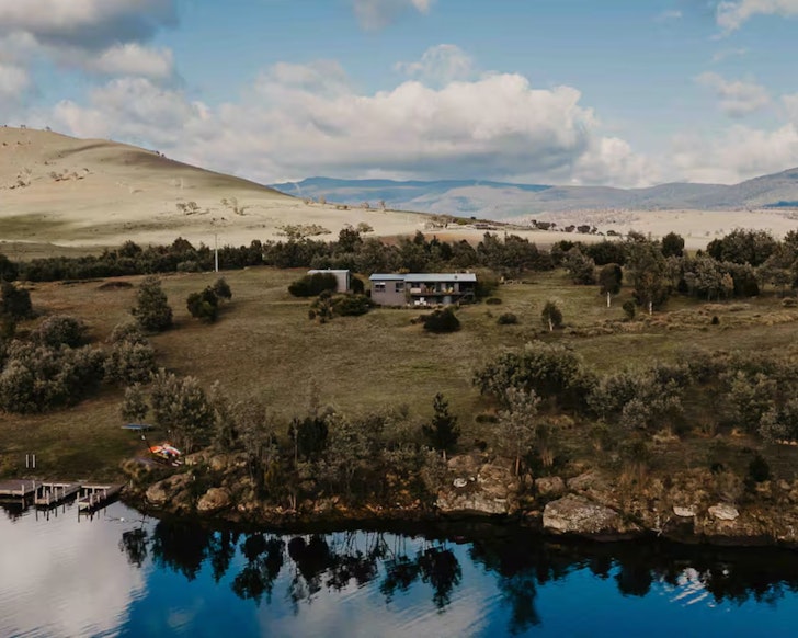 A view of The Lake House surrounded by fields and with the lake in front in Tasmania