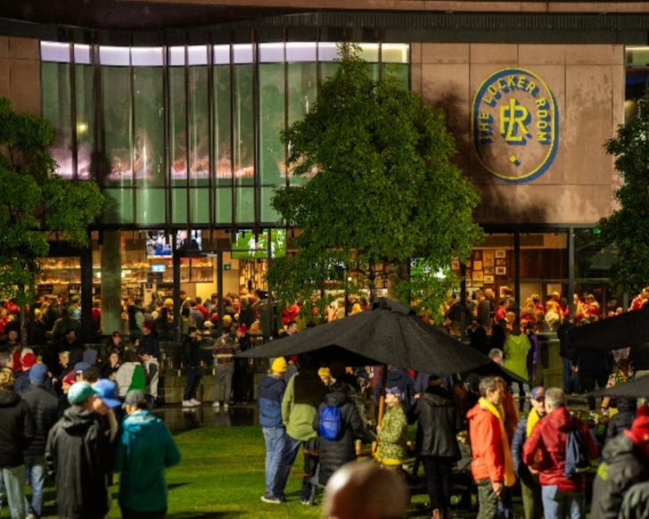Outdoor wide shot of a crowd of people mingling and drinking outside of The Locker Room building. TV screens playing live sport are visible through the windows, and the picture is taken at night time.