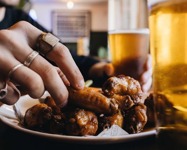 Someone with interesting rings leans in to pick up a chicken wing off a plate.