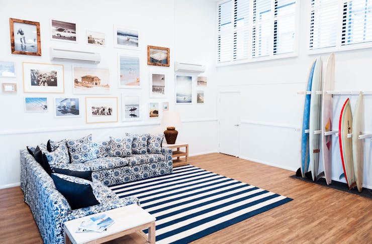 A blue and white L-shape couch sits underneath a gallery wall at a Byron Bay hostel. There is a rack of surfboards against the wall.