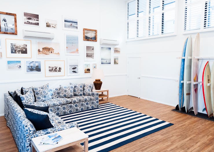 A blue and white L-shape couch sits underneath a gallery wall at a Byron Bay hostel. There is a rack of surfboards against the wall.