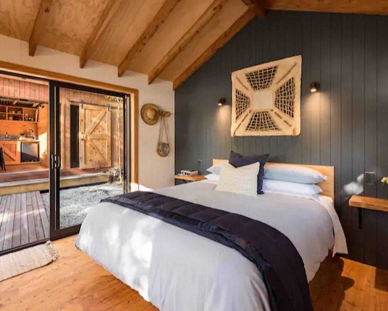 A large, cosy bed sits in a wooden cabin. In the background we see another cabin, housing a kitchen. 