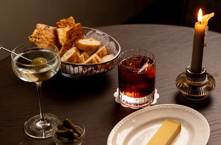 A basket of bread, a slab of cheese, a negroni and a single lit candle on a table. 